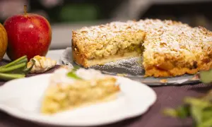 Krucha szarlotka z kawałkami kwaśnych jabłek, posypana cukrem pudrem. Obok leżą świeże jabłka i mały rogalik.