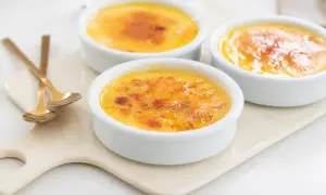 Tre krämiga creme brulee i vita skålar med karamelliserat sockerlock. Guldfärgade skedar ligger bredvid.