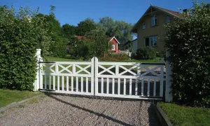 Vit grind med korsmönster leder in på en grusgång entre till ett gult hus och en röd stuga.
