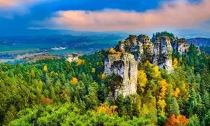 Felsformationen im böhmischen Paradies (Český ráj) ragen aus einem bunten Herbstwald hervor, unter einem dramatischen Himmel.