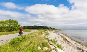 En cyklist njuter av en vacker dag längs en kustväg, en av många cykelleder i Danmark. Havet glittrar bredvid den steniga stranden.