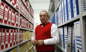 Starszy mężczyzna w czerwonym swetrze stoi między regałami pełnymi segregatorów.