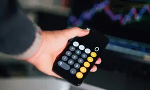 Dłoń trzyma smartfon z kalkulatorem, na ekranie widać wykresy finansowe. Obliczanie faktury bez vat.