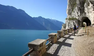Ciclisti sul sentiero della Ponale, con il lago e le montagne sullo sfondo. Un tunnel scavato nella roccia attende.