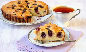 Una fetta di torta bertolina, soffice e profumata, con ciliegie mature, accanto a una tazza di tè fumante.