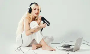 Elena Rose, a blonde woman with tattoos, sits on the floor wearing headphones and holding a microphone, ready to record.