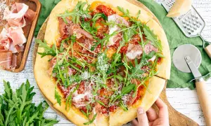Pizza mit Rucola, Schinken und Parmesan, daneben Prosciutto, Parmesan und Pizzaschneider.