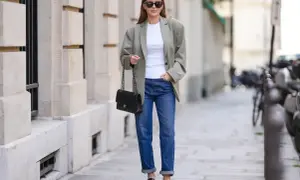 Kobieta w luźnym, beżowym blazerze, białej koszulce i jeansach prezentuje casual damski przykłady. Czarna torebka na łańcuszku i mokasyny dopełniają stylizację.