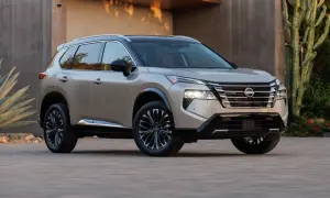 Elegancki, beżowy Nissan Rogue z czarnym dachem i przyciemnianymi felgami stoi przed nowoczesnym budynkiem, otoczony egzotyczną roślinnością.