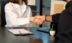 Uścisk dłoni po ustaleniu formy prawnej podmiotu. Na stole dokumenty, laptop i roślina.