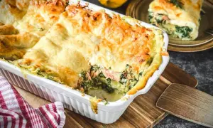 Herzhafte Spinat-Lachs-Lasagne, goldbraun überbacken, dampft in einer weißen Auflaufform. Ein Stück auf einem Teller daneben zeigt die Schichten.