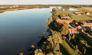 Spokojne jezioro Dejguny otoczone zielenią i domkami z czerwonymi dachami. W oddali widać lasy i pola.
