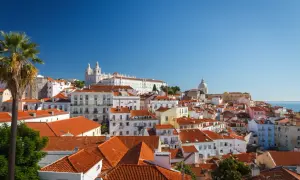 Blick über die Dächer der Lissabon Altstadt mit Palmen und Kirchen im Hintergrund unter blauem Himmel.