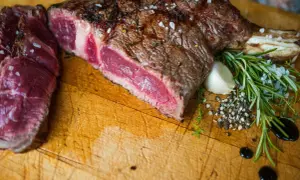 Une belle côte de bœuf saignante, assaisonnée de gros sel et de poivre, avec du romarin et de l'ail sur une planche en bois.