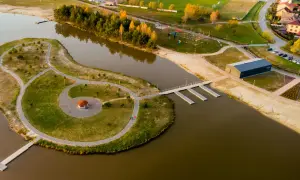 Malownicze, nisko jezioro z wyspą, ścieżkami, altanką i nowoczesnym budynkiem. W tle widać drzewa, plażę i zabudowania.