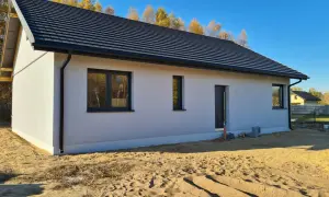 Nowy dom z szarym dachem i białymi ścianami, otoczony piaskiem i jesiennymi drzewami. Ssz.