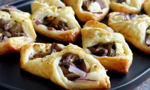 Chrupiące ciasto francuskie z pieczarkami, serem feta i czerwoną cebulą, ułożone na czarnej tacy.
