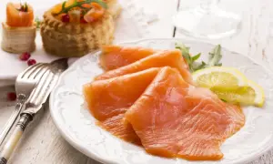 Tranches de saumon fumé, garnies de roquette et de citron, sur une assiette blanche. Des bouchées feuilletées au saumon sont visibles en arrière-plan.