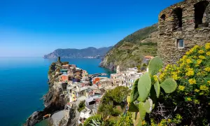 Kolorowe domy Cinque Terre wznoszą się na klifie nad turkusowym morzem. W oddali widać góry, a na pierwszym planie kaktus i żółte kwiaty.