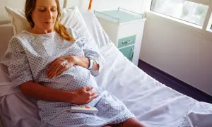 Kobieta w szpitalnym łóżku, z kroplówką i opaską na ręce, czeka na dziecko. Jej tryb życia po założeniu szwu szyjkowego jest spokojny.