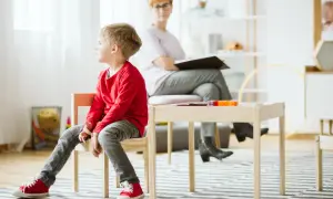 Pojke i röd tröja sitter på en stol under en autism utredning barn. En kvinna sitter bakom ett bord.