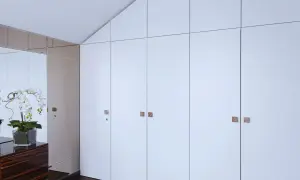 Armario empotrado con puertas blancas y tiradores cuadrados. Reflejos de orquídeas y un mueble de madera oscura en el espejo. Ideal para cambiar puertas armario empotrado.
