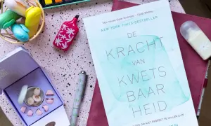 Boek 'De Kracht van Kwetsbaarheid' en oordopjes, ideaal voor wie last heeft van omgevingsgeluid. Creatieve spullen op een roze ondergrond.