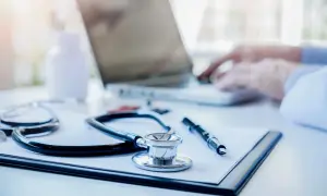Stetoskop, długopis i notatnik na biurku lekarskim, w tle widać laptop i dłonie piszące na klawiaturze.