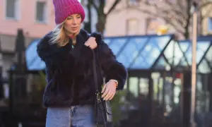 Krótkie futerko stylizacje: blondynka w różowej czapce i jeansach, z czarną torebką.