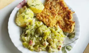 Kotlet schabowy z ziemniakami i zasmażaną kapustą z boczkiem na talerzu.