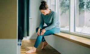 Femme assise près d'une fenêtre, tenant son genou, possiblement à cause d'une **durée courbature** après un effort.