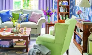 Jasny salon z fioletowymi zasłonami i kwiecistą tapetą. Wygodna kanapa i zielony fotel tworzą przytulną atmosferę, idealną na wiosnę w domu.