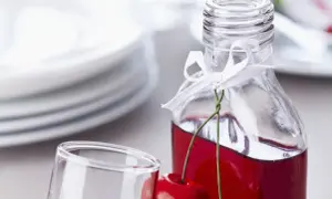 Szklana butelka z nalewką wiśniową, ozdobiona wstążką i wiśniami, obok kieliszka.