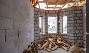 Interno di una casa in costruzione con muri in blocchi di cemento, travi in legno a vista e cumuli di legname. Sembra un cantiere fermo da tempo, quasi un abuso edilizio dopo 20 anni.