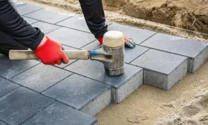 Ręce w czerwonych rękawiczkach układają szare kostki brukowe, tworząc ścieżki. Młotek gumowy dociska ostatnią kostkę.