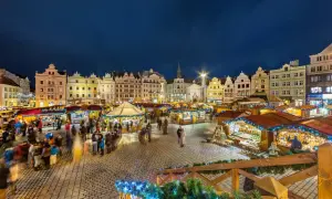 Beleuchtete Buden und historische Gebäude auf einem tschechischen Weihnachtsmarkt. Menschen schlendern über den Platz, der mit Lichterketten geschmückt ist.