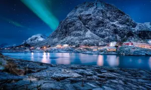 Grüne Bänder der Aurora Borealis tanzen über einem verschneiten Berg, der eine kleine Küstensiedlung mit roten und gelben Häusern beleuchtet.