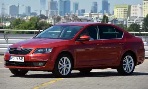 Czerwona Skoda Octavia 3 z widocznymi żarówkami w przednich reflektorach, zaparkowana na tle miasta.