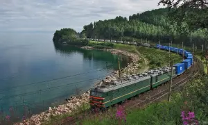 Zielony pociąg towarowy jedzie wzdłuż malowniczego jeziora. To fragment trasy kolej transsyberyjska, gdzie tory biegną blisko wody, otoczone zielenią i skalistym brzegiem.