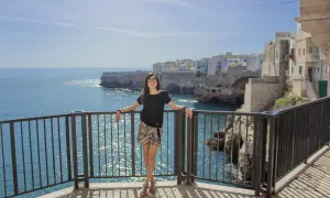 Donna sorridente con occhiali da sole si appoggia a una ringhiera con vista sul mare cristallino e le case bianche di Polignano a mare.