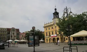 Rynek w Ostrowie Wielkopolskim z żółtym ratuszem i wieżą zegarową. W tle kamienice, drzewa i ogródek kawiarniany.