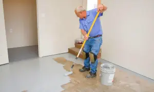 Un homme en bleu peint le sol en béton avec un rouleau. Il porte des genouillères pour peindre le béton confortablement.