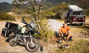 Motocykl, namiot, kamper i mężczyzna siedzący przy ognisku na tle gór.