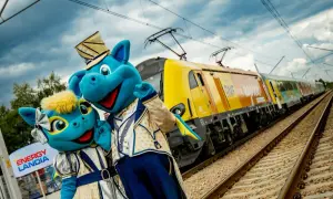 Dwa niebieskie smoki w strojach stoją obok żółtego pociągu na tle torów i nieba.
