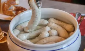 Eine Gabel hebt eine Weißwurst aus einem Topf, um sie zuzubereiten. Daneben liegt eine Brezel.