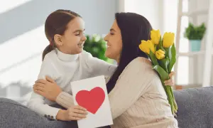 Uśmiechnięta dziewczynka wręcza mamie kartkę z sercem i bukiet żółtych tulipanów. Radosny Dzień Matki UK!