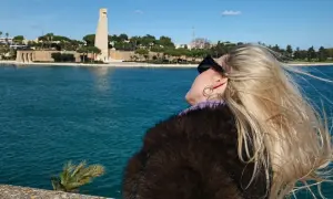 Kobieta z blond włosami podziwia pomnik w Brindisi. W tle błękitne morze i błękitne niebo.
