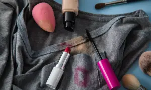 Graues T-Shirt mit Flecken von Lippenstift, Mascara und Foundation. Ein Make-up-Schwamm und Pinsel liegen daneben. Tipps zum Make-up aus Kleidung entfernen.