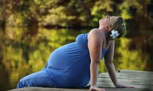 Gravid kvinna i blå klänning på en brygga, med en blomma i håret. Hon lutar sig bakåt, kanske känner hon av Braxton Hicks-sammandragningar.
