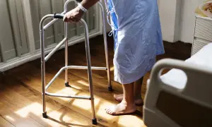 Osoba w szpitalnej koszuli, wspierając się na chodziku, stawia pierwsze kroki po udarze.